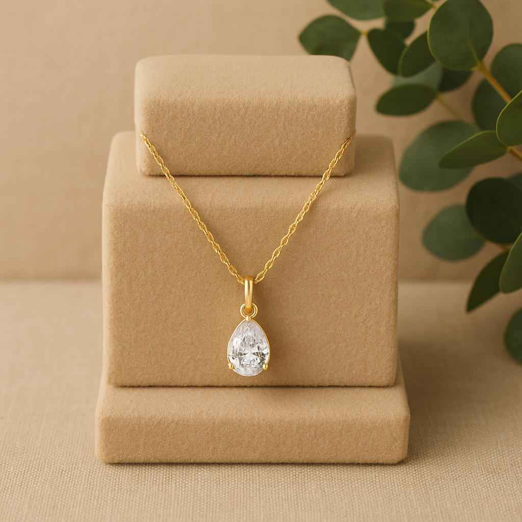 Gold necklace with a teardrop-shaped pendant on a beige stand with a green leafy background