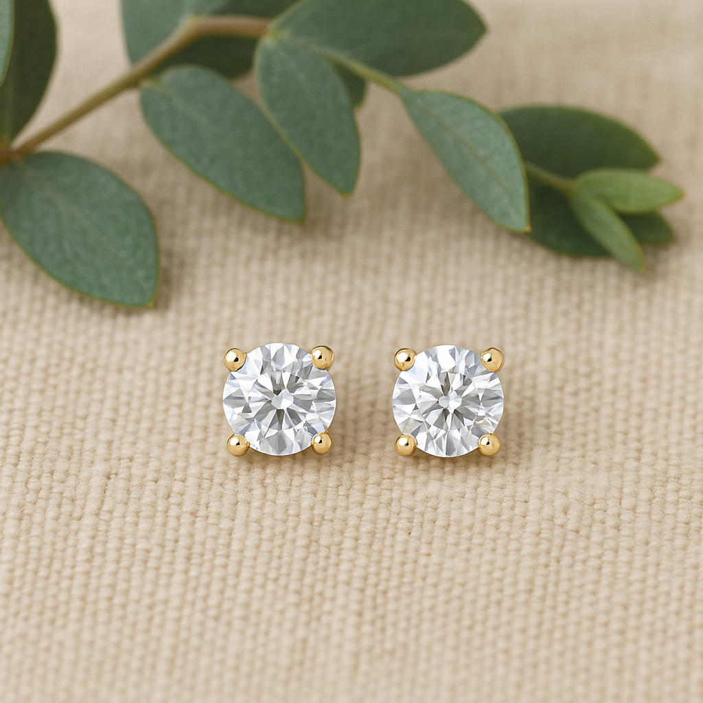 Gold stud earrings with clear gemstones on a beige fabric background with green leaves.